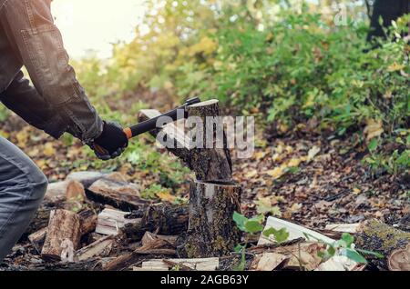 Man schneidet ein Protokoll mit einem industriellen Ax. Teile des Baumes fliegen auseinander. Ein starker Mann hält eine Axt in der Hand auf einem Hintergrund von Wald, Brennholz. Ein Mann lu Stockfoto