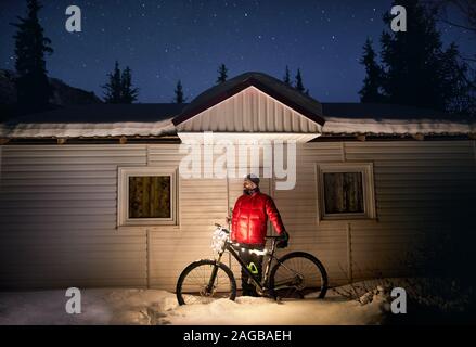 Mann im roten Mantel mit Fahrrad mit Weihnachtsbeleuchtung in der Nähe von kleines Haus im Winter verschneite Wald in die Berge unter dem Nachthimmel mit Sternen