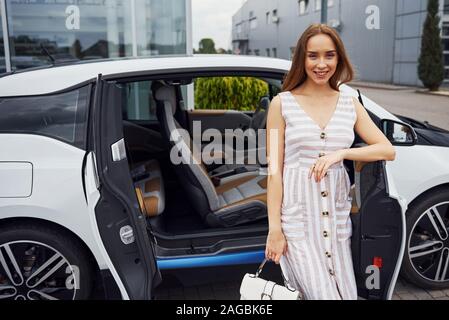 Mit Tasche in der Hand. Frau mit blonden Haaren steht in der Nähe von Ihrem weißen Auto auf der Straße Stockfoto