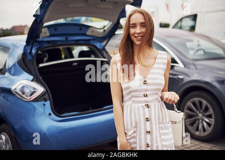 Frau mit blonden Haaren steht in der Nähe von ihren blauen Auto auf der Straße mit Tasche in der Hand Stockfoto