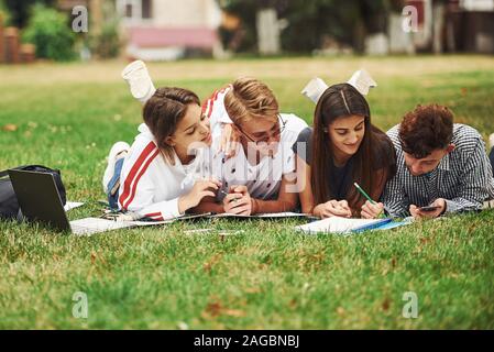 Schreiben auf Papier. Vorderansicht. Gruppe von jungen Studenten in Freizeitkleidung auf grünem Gras tagsüber Stockfoto