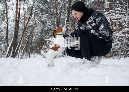 Von der Seite. Mann in warme Kleidung sitzen und spielen mit seinem Hund im Winter Forest Stockfoto