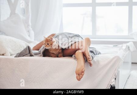 Beine der schlafende Mädchen auf dem weißen Bett. Süße Katze ist wach und will irgendwo gehen Stockfoto