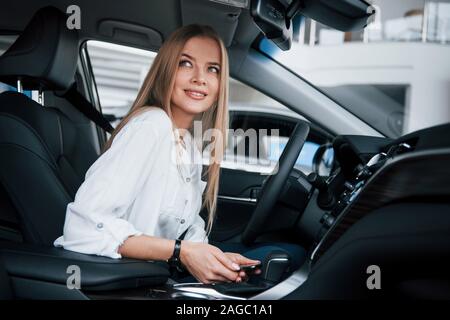 Hält Smartphone und lächelnd. Schöne blonde Mädchen in der neuen Auto sitzen mit modernen schwarzen Interieur Stockfoto