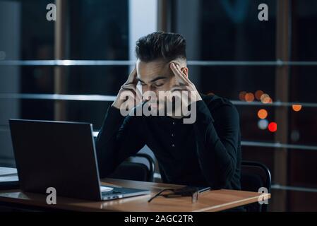 Starke Kopfschmerzen. Stilvolle Junge Unternehmer arbeitet allein im Büro bei Nacht Stockfoto