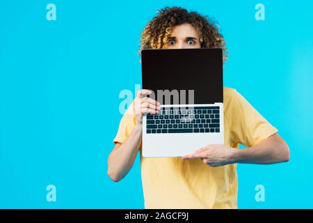Erschrocken curly Student versteckt sich hinter Laptop mit leerer Bildschirm isoliert auf Blau Stockfoto