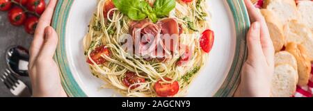 Selektiver Fokus der Frau mit Platte mit Pappardelle mit Tomaten, Basilikum und Prosciutto in der Nähe von Baguette, Tomaten und Besteck auf graue Oberfläche, panor Stockfoto