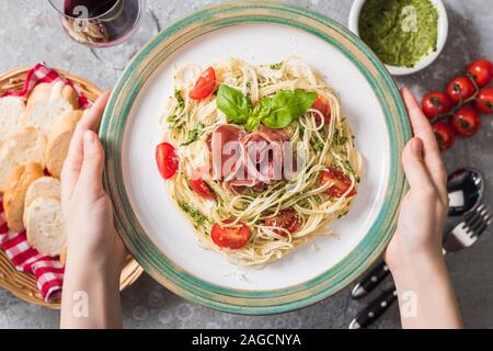 Selektiver Fokus der Frau mit Platte mit Pappardelle mit Tomaten, Basilikum und Prosciutto in der Nähe von Baguette, Rotwein, Pesto, Tomaten und Besteck auf gr Stockfoto