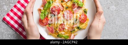 Teilweise mit Blick auf die Frau mit Platte mit Pappardelle mit Tomaten, Basilikum und Prosciutto auf plaid Serviette auf graue Oberfläche, Panorama-Aufnahme ein. Stockfoto