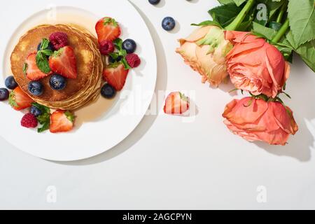 Blick von oben auf die leckere Pfannkuchen mit Honig, Blaubeeren und Erdbeeren auf Platte in der Nähe der Rosen auf Marmor weiß Oberfläche Stockfoto