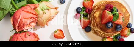 Blick von oben auf die leckere Pfannkuchen mit Honig, Blaubeeren und Erdbeeren auf Platte in der Nähe der Rosen auf Marmor weiß Oberfläche, Panorama-Aufnahme ein. Stockfoto