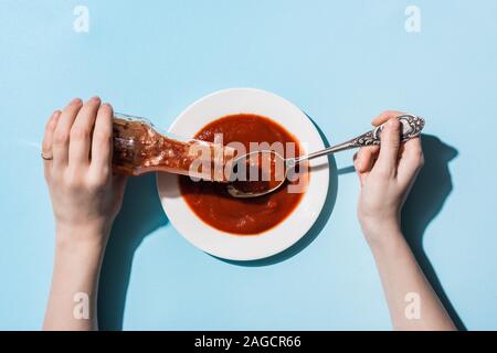 7/8-Ansicht von Frau gießen Ketchup aus der Flasche auf dem Teller auf blauem Hintergrund Stockfoto