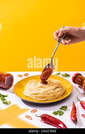 7/8-Ansicht von Frau gießen Ketchup auf Spaghetti neben Chili und Koriander auf weisse Oberfläche isoliert auf Gelb Stockfoto