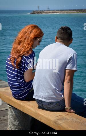 Eine Ansicht der Rückseite eine rothaarige junge Frau mit langen Haaren neben einem jungen Mann auf einer Holzbank am Pier mit Blick auf den Pazifischen Ozean sitzen Stockfoto