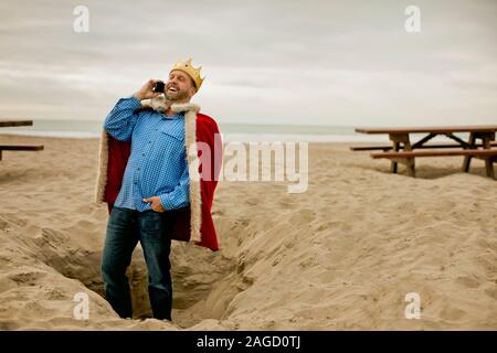 Portrait von Lachen Mitte - erwachsener Mann mit Krone und Robe, während sein Handy am Strand. Stockfoto