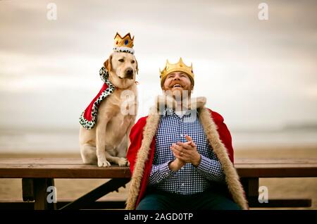 Mitte - erwachsener Mann mit Mantel und Krone und Lachen beim Sitzen auf der Bank mit seinem Hund am Strand. Stockfoto