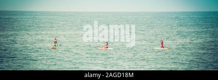 Ein Panoramablick auf das Format einer Ansicht der Rückseite zwei Bikini bekleideten Frauen und ein Mann kniet, wie sie ihren Handlungsspielraum Stand up Paddle Boards aus in Richtung Pazifik Stockfoto