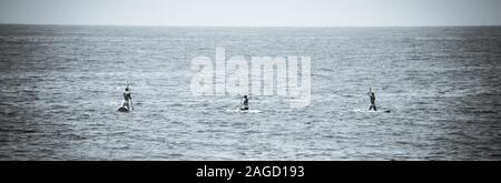 Ein Panoramablick auf das Format einer Ansicht der Rückseite zwei Bikini bekleideten Frauen und ein Mann kniet, wie sie ihren Handlungsspielraum Stand up Paddle Boards aus in Richtung Pazifik Stockfoto