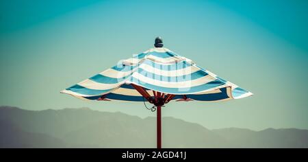 Ein 7/8-panoramaformat Sicht auf den Himmel und ein einsames blau gestreifter Sonnenschirm mit atmosphärischen Foggy Mountain Range Hintergrund in Südkalifornien Stockfoto