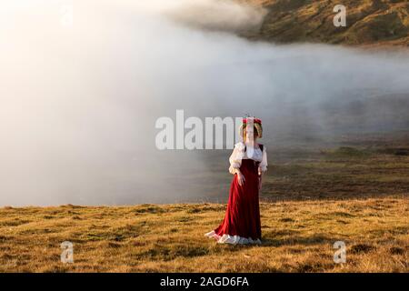 Eine junge rothaarige Frau in altmodischer Kleidung mit einem leuchtend roten Rock bleibt auf dem Feld im Hochland. Faröer Island. Ich habe eine Modellversion Stockfoto