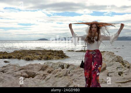 Hippie modische junge Dame mit langen braunen Haaren und floralen Schlaghosen am Meer auf Felsen unter einem cloudscape posiert, schmollen und Spielen wit Stockfoto