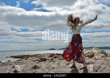 Hippie modische junge Dame mit langen braunen Haaren und floralen Schlaghosen posing und Tanz am Meer auf Felsen unter einem CLOUDSCAPE, Waffen in den Stockfoto