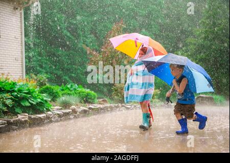 Zwei kleine Kinder mit Regenschirmen und in Gummistiefeln spielen Draußen während des Regens Stockfoto