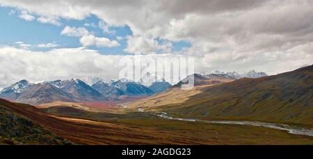 Panorama-Landschaft von hohen felsigen Bergen im Schnee bedeckt Umgeben von viel Grün und einem Fluss Stockfoto