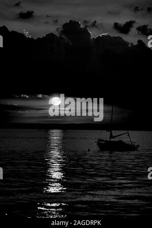 Graustufenaufnahme eines Schiffes im Meer unter dem Dunkler Wolkenhimmel mit dem Mond im Hintergrund Stockfoto