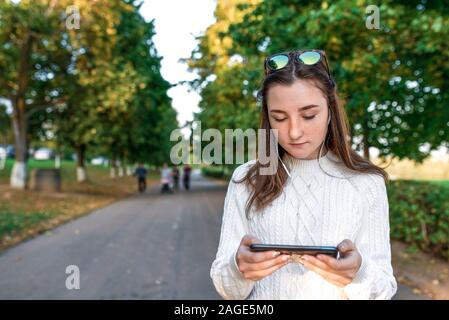 Jugendlicher Mädchen Uhren Video auf Handy, Sommer Park, Hintergrund Herbst Bäume, hört Musik mit Kopfhörer, online im Internet, gratis Stockfoto