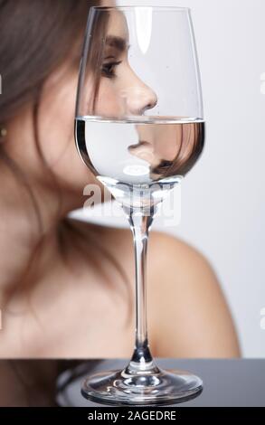 Mädchen verbirgt ihr Gesicht hinter einem Glas Glas Wasser. Schönheit Porträt der jungen Frau. Weibliche auf grauem Hintergrund. Stockfoto