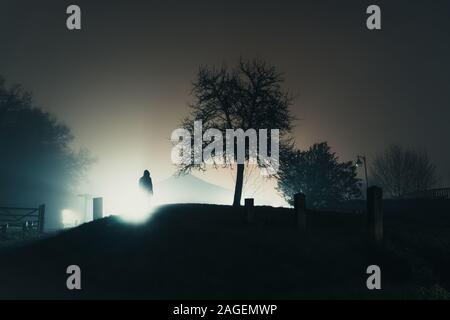 Eine vermummte Gestalt gegen Straßenlaternen Silhouette, die neben einem Baum. Auf einer geheimnisvollen Nebel, winter nacht Stockfoto