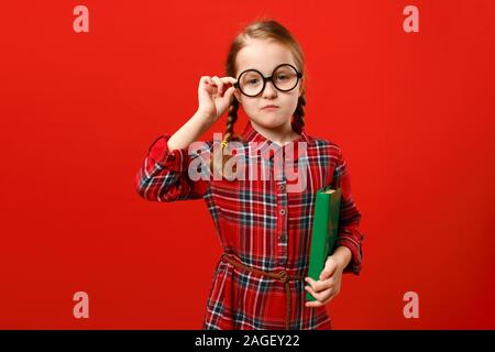 Lustig Kind in Gläsern und mit einem Buch auf einem roten Hintergrund. Porträt einer ernsten Smart kleines Mädchen. Stockfoto