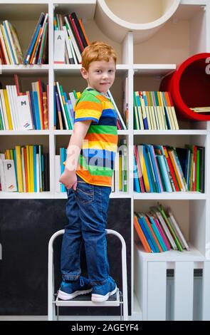 Adorable rothaarige Kleine Junge, stehend auf einer Treppe in der Bibliothek. Childs Gehirn Entwicklung, Lernen, Lesen, kognitive Fähigkeiten. Fertig für die Schule. Ch Stockfoto