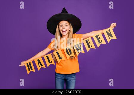 Foto von kleine Hexe Dame spielen paranormale Wesen Thema halloween party Holding feier Fahnen tragen Orange t-shirt wizard Hut isoliert Lila Stockfoto