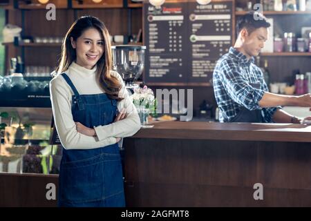 Portrait von jungen erwachsenen Asiatischen Inhaber der Cafeteria Cafe vor Barista Bar im Coffee Shop mit einem Mitarbeiter im Hintergrund arbeiten. Verwendung für e Stockfoto