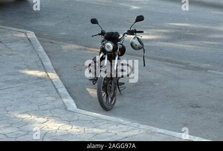 Eine alte schwarze Motorrad auf der Seite einer Straße geparkt werden. Es hat einen Silbernen Helm hängen von der Lenker. Stockfoto