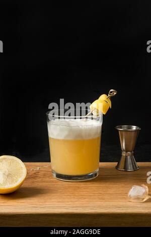 Whiskey Sour Cocktail - Bourbon mit Zitronensaft, Zucker, Sirup und Eiweiß in Glas auf Holzbrett auf Schwarz. Vertikale Ausrichtung. Stockfoto