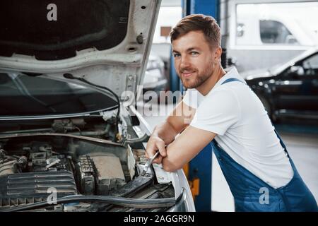 Positiver Mann lehnte sich auf das Auto. Mitarbeiter in der blauen Uniform arbeitet im Automobil Salon Stockfoto