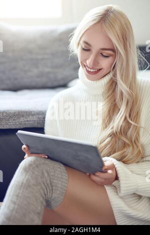 Trendy lächelnde blonde Frau im Winter Leggins und einem warmen Wollpullover zu Hause entspannen mit einem Tablet-PC lesen mit einem schönen lebhaften Lächeln Stockfoto