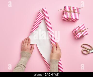 Zwei weibliche Hände eine Rolle Geschenkpapier, neben zwei Geschenke, rosa Hintergrund entfalten, Überraschungen vorbereiten Stockfoto