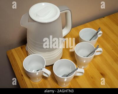 Weißer Wasserkocher und vier weiße Keramik Becher mit Löffeln auf einem Holztisch Stockfoto