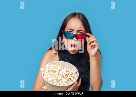 Freestyle. Junge Frau mit 3D-Brille isoliert auf Blau spuckt Popcorn entrüstet stehend Stockfoto