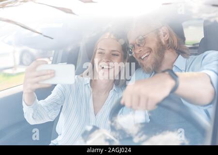 Transport. Junges Paar mit dem Elektroauto Freund fahren während die Freundin, die auf dem Smartphone selfie posiert für die Kamera fröhliche anzeigen Stockfoto