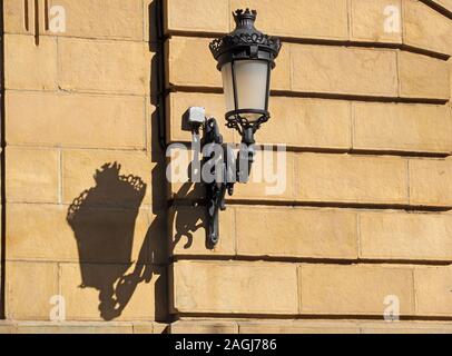 Aufwändige schwarzen schmiedeeisernen Straßenlaterne und Halterung wirft einen Schatten auf die Diagonale parallel Kurse von Gekleideten Steinmauer in San Sebastian, Spanien, Europa Stockfoto
