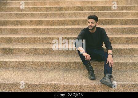 Junge schöne Inder denken und suchen im Abstand beim Sitzen auf der Treppe aus Granit texturierte Treppe Stockfoto