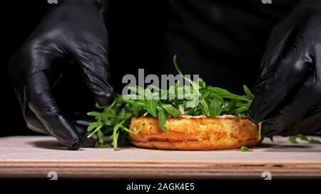 Handwerk Burger ist Kochen auf schwarzem Hintergrund. Bestehen: sauce, Rucola, Tomaten, roten Zwiebeln Pommes, Gurken, Käse, Brötchen und Marmor Fleisch Rindfleisch. Stockfoto