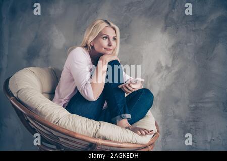 Voller Körper Foto des friedlichen verträumte Frau, die ihr Handy schauen Sie haben Träume Pläne über ihre Wochenenden sitzen in weichen, weißen Sessel im Wohnzimmer Stockfoto