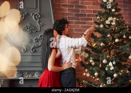 Die Vorbereitung für den Urlaub. Romantisches Paar Dressing up Weihnachtsbaum im Zimmer mit braunen Wand und Kamin Stockfoto