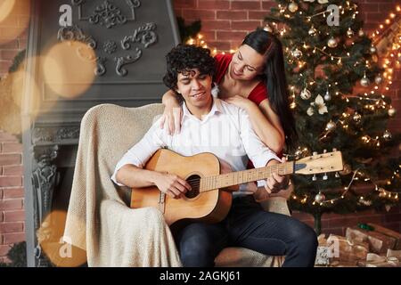 Lockigen Haaren attraktiver Kerl auf dem Stuhl mit der akustischen Gitarre mit Weihnachtsbaum hinter sich. Freundin in weißem Kleid ist umarmt ihren Freund Stockfoto